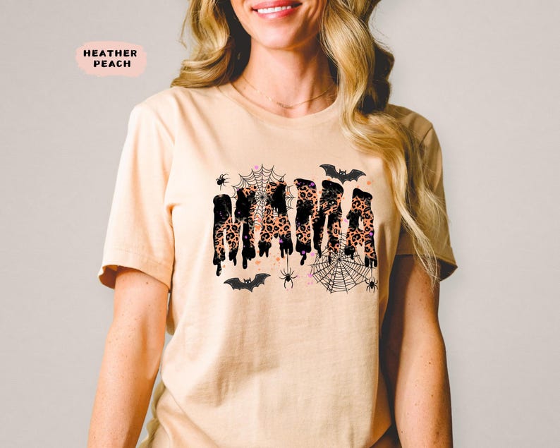 May include: Heather peach-colored t-shirt with the word "MAMA" in a dripping, leopard print design. The shirt features spiderwebs, spiders, and bats, creating a Halloween theme. The shirt is a short-sleeved crew neck.