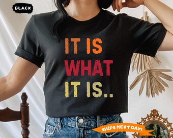 May include: Black t-shirt with the phrase "IT IS WHAT IT IS" in orange, red, and yellow block letters. The shirt has short sleeves and a crew neck. The word "BLACK" is in a black box in the upper left corner.