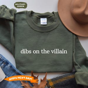 May include: Military green sweatshirt with the text "dibs on the villain" in white. The sweatshirt is displayed with a brown hat, jeans, and a coffee cup. The sweatshirt is a crew neck style.
