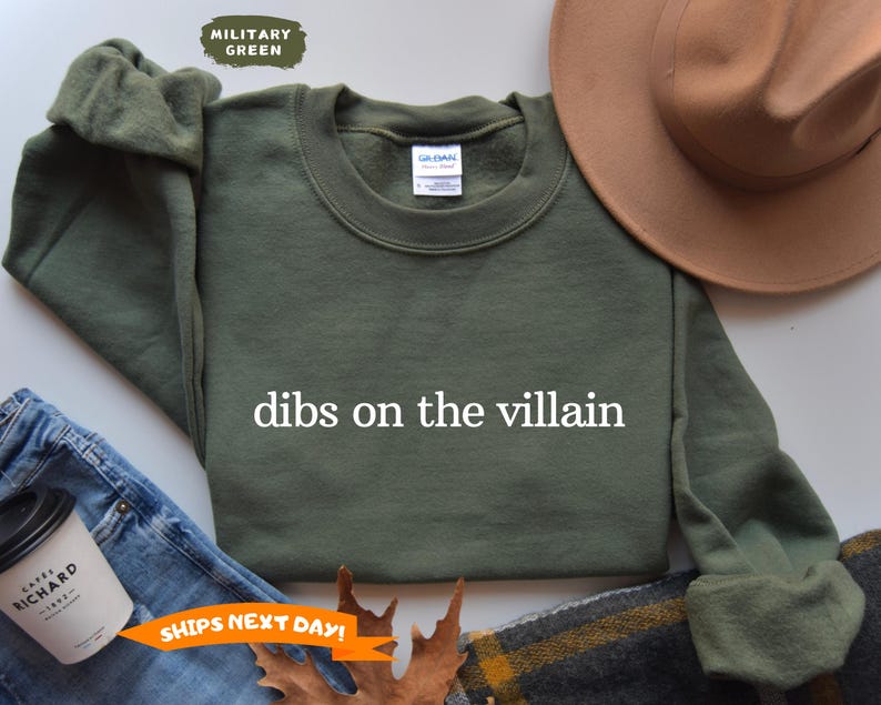 May include: Military green sweatshirt with the text "dibs on the villain" in white. The sweatshirt is displayed with a brown hat, jeans, and a coffee cup. The sweatshirt is a crew neck style.