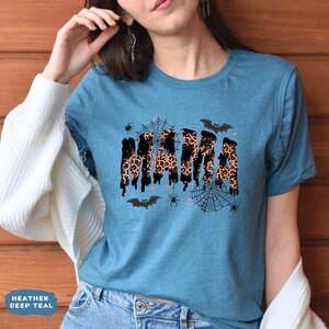 May include: A teal t-shirt with the word "MAMA" in a leopard print design, dripping with black accents. The shirt features Halloween-themed graphics, including bats, spiders, and spiderwebs. The text "Heather Deep Teal" is at the bottom left.