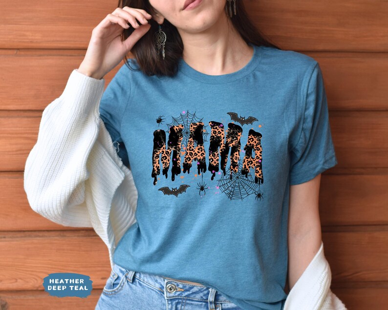May include: A teal t-shirt with the word "MAMA" in a leopard print design, dripping with black accents. The shirt features Halloween-themed graphics, including bats, spiders, and spiderwebs. The text "Heather Deep Teal" is at the bottom left.