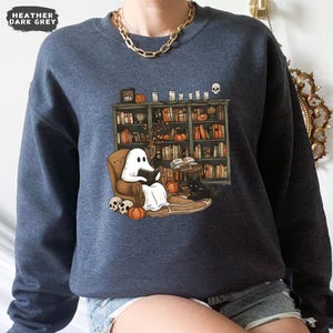 May include: Heather dark gray sweatshirt featuring a whimsical illustration of a ghost reading in a cozy armchair in front of a bookshelf. Pumpkins, candles, and a black cat add to the Halloween theme.
