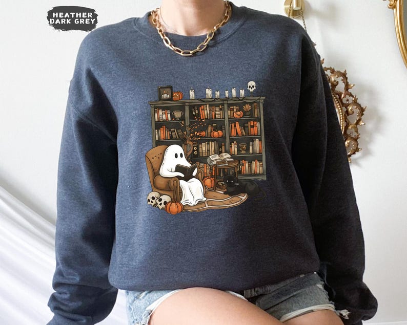 May include: Heather dark gray sweatshirt featuring a whimsical illustration of a ghost reading in a cozy armchair in front of a bookshelf. Pumpkins, candles, and a black cat add to the Halloween theme.