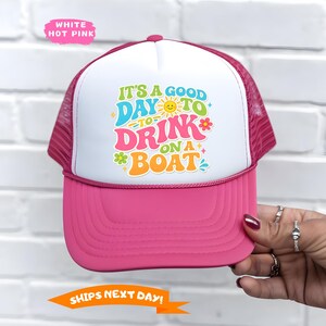 May include: A hot pink and white trucker hat with the text "IT'S A GOOD DAY TO DRINK ON A BOAT" in colorful letters. The hat has a mesh back and a curved brim. The hat is a fun accessory for boating or outdoor activities.