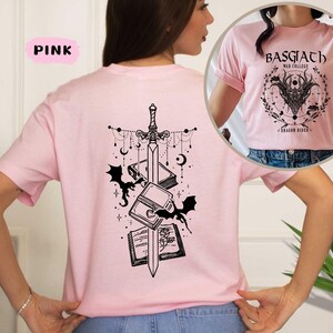 May include: Pink t-shirt with a black graphic design on the back featuring a sword, books, bats, and celestial elements. The front of the shirt has a circular design with the text "BASGIATH WAR COLLEGE" and a dragon illustration. The shirt is a light pink color.