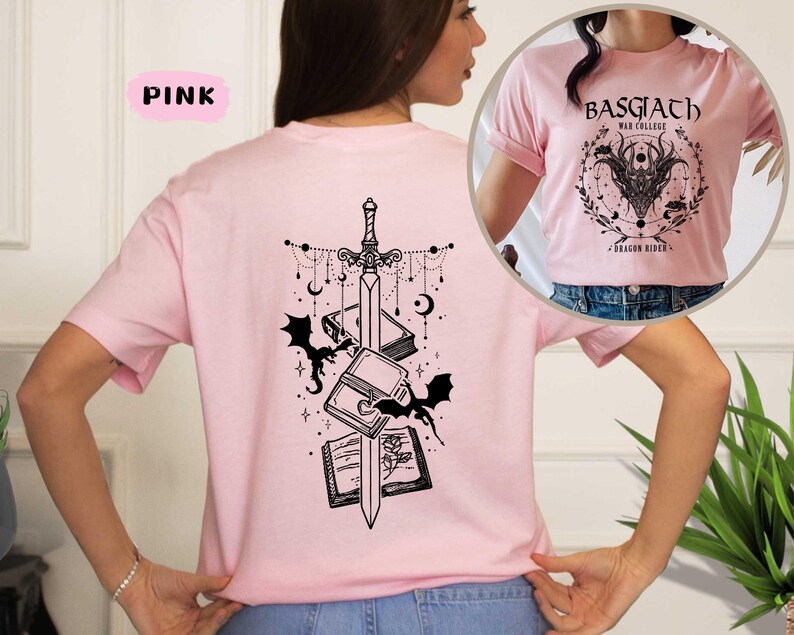 May include: Pink t-shirt with a black graphic design on the back featuring a sword, books, bats, and celestial elements. The front of the shirt has a circular design with the text "BASGIATH WAR COLLEGE" and a dragon illustration. The shirt is a light pink color.