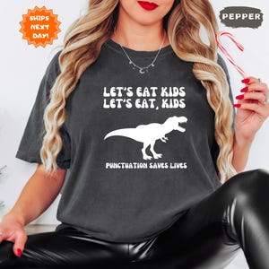 May include: Dark gray t-shirt with the text "LET'S EAT KIDS, LET'S EAT, KIDS" and a white dinosaur graphic. The phrase "PUNCTUATION SAVES LIVES" is below the dinosaur. The shirt is a comfortable fit.