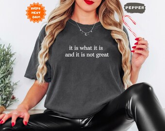 May include: Dark gray t-shirt with the text "it is what it is and it is not great" in white. The person is wearing black leather pants and holding a candy cane. A small green plant is on the left.
