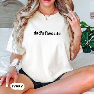 May include: Ivory-colored t-shirt with the text "dad's favorite" in black. The shirt is a relaxed fit and appears to be made of a soft material. The word "IVORY" is written on a small label.