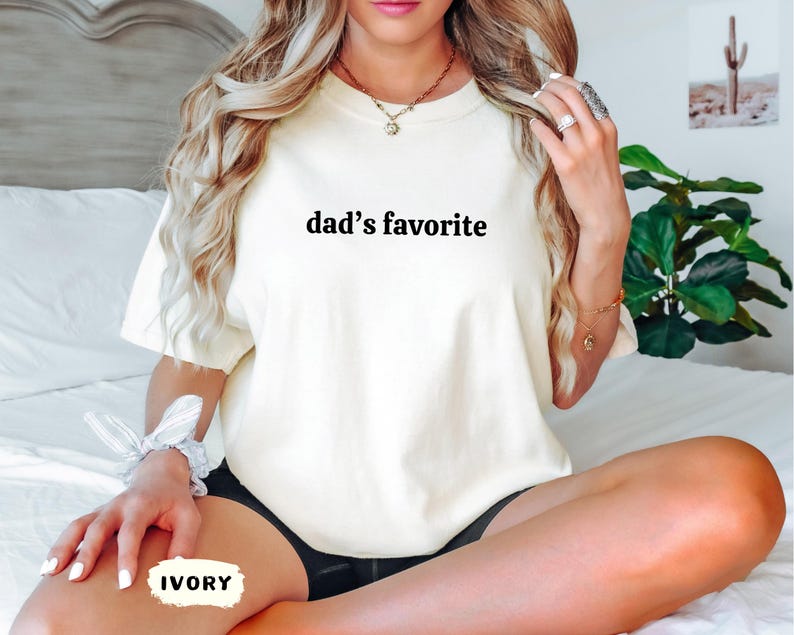 May include: Ivory-colored t-shirt with the text "dad's favorite" in black. The shirt is a relaxed fit and appears to be made of a soft material. The word "IVORY" is written on a small label.
