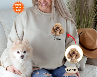 May include: A beige sweatshirt with a personalized design featuring a dog's face and the name "Cooper". The sweatshirt has a pocket on the front and the word "SAND" printed on the bottom right corner.