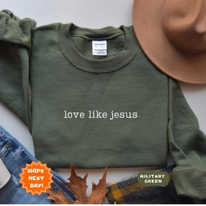 May include: An olive green sweatshirt with the text "love like jesus" in white. A brown hat, blue jeans, and a coffee cup are also in the image. The sweatshirt is labeled "Military Green".