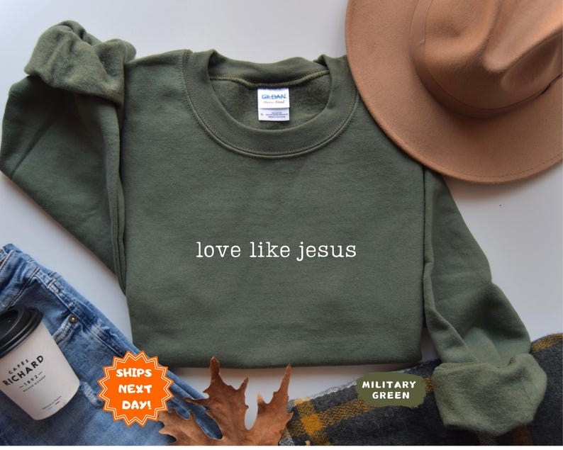 May include: An olive green sweatshirt with the text "love like jesus" in white. A brown hat, blue jeans, and a coffee cup are also in the image. The sweatshirt is labeled "Military Green".