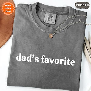 May include: A gray Comfort Colors t-shirt with the text "dad's favorite" in white. The shirt has a crew neck and short sleeves. A gold necklace with a pendant is draped across the shirt. A brown hat and dried leaves are in the background.