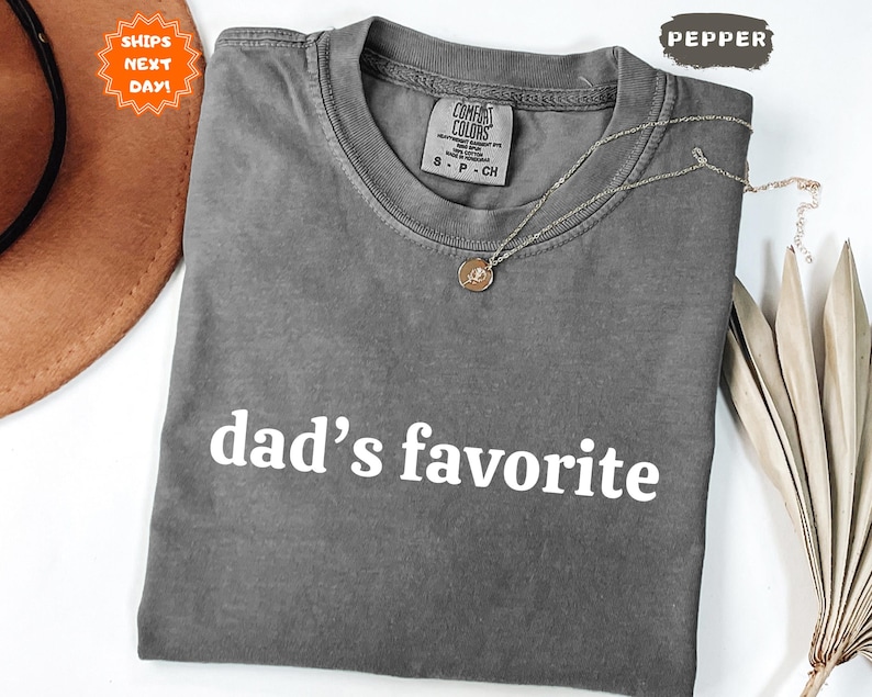 May include: A gray Comfort Colors t-shirt with the text "dad's favorite" in white. The shirt has a crew neck and short sleeves. A gold necklace with a pendant is draped across the shirt. A brown hat and dried leaves are in the background.