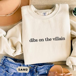 May include: A cream-coloured sweatshirt with the text "dibs on the villain" printed in black. The sweatshirt is on a wooden surface with blue jeans, a brown hat, and sandals.
