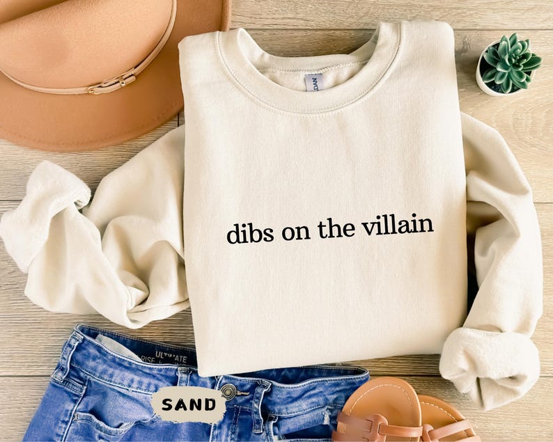 May include: A cream-coloured sweatshirt with the text "dibs on the villain" printed in black. The sweatshirt is on a wooden surface with blue jeans, a brown hat, and sandals.