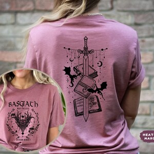 May include: Heather maroon t-shirt with a black graphic on the back featuring a sword, books, bats, and celestial elements. The front of the shirt has a circular design with text and a dragon illustration. The shirt is a crew neck.