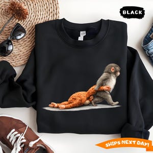 May include: Black crewneck sweatshirt featuring a graphic of a baby monkey carrying a stuffed animal. The sweatshirt has a classic fit and is made of a soft material. The word "BLACK" is printed on the top right corner.