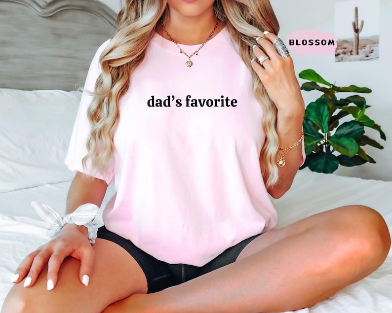 May include: A light pink t-shirt with the text "dad's favorite" in black. The shirt is a crew neck style and appears to be made of a soft material. The model is wearing black shorts.