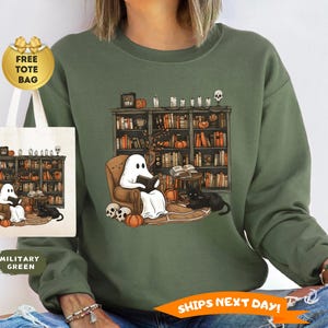 May include: Military green sweatshirt featuring a Halloween-themed graphic. The design shows a ghost reading in a cozy armchair in front of a bookshelf, with a black cat, pumpkins, and skulls. A tote bag with the same design is included.