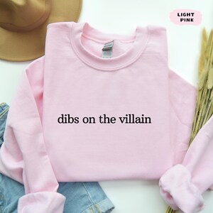 May include: Pale pink crew neck sweatshirt with the text "dibs on the villain" in black. The sweatshirt is folded. A brown hat and blue denim jeans are partially visible in the background.