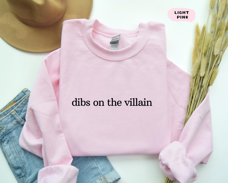 May include: Pale pink crew neck sweatshirt with the text "dibs on the villain" in black. The sweatshirt is folded. A brown hat and blue denim jeans are partially visible in the background.