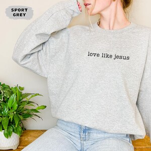 May include: A light gray sweatshirt with the text "love like jesus" in black. The sweatshirt is a sport grey color and has long sleeves. The image also shows a green plant in a white pot.