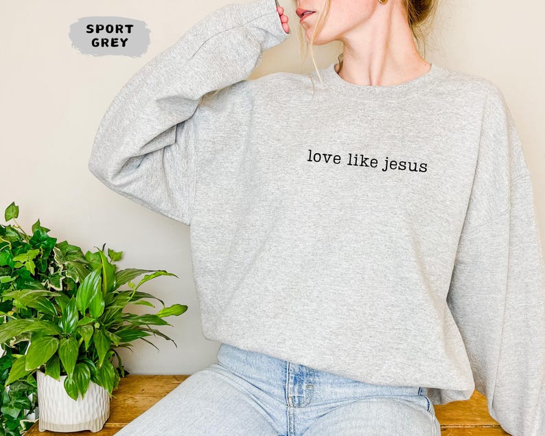 May include: A light gray sweatshirt with the text "love like jesus" in black. The sweatshirt is a sport grey color and has long sleeves. The image also shows a green plant in a white pot.