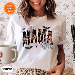 May include: White t-shirt with a Halloween-themed design. The word "MAMA" is printed in a dripping style with a leopard print pattern. The design includes bats, spiders, and spiderwebs.