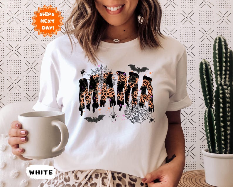 May include: White t-shirt with a Halloween-themed design. The word "MAMA" is printed in a dripping style with a leopard print pattern. The design includes bats, spiders, and spiderwebs.