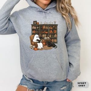 May include: A light gray hoodie featuring a Halloween-themed graphic. The graphic depicts a ghost reading in a chair in front of a bookshelf, with pumpkins, a black cat, and skulls. The text "SPORT GREY" is also visible.