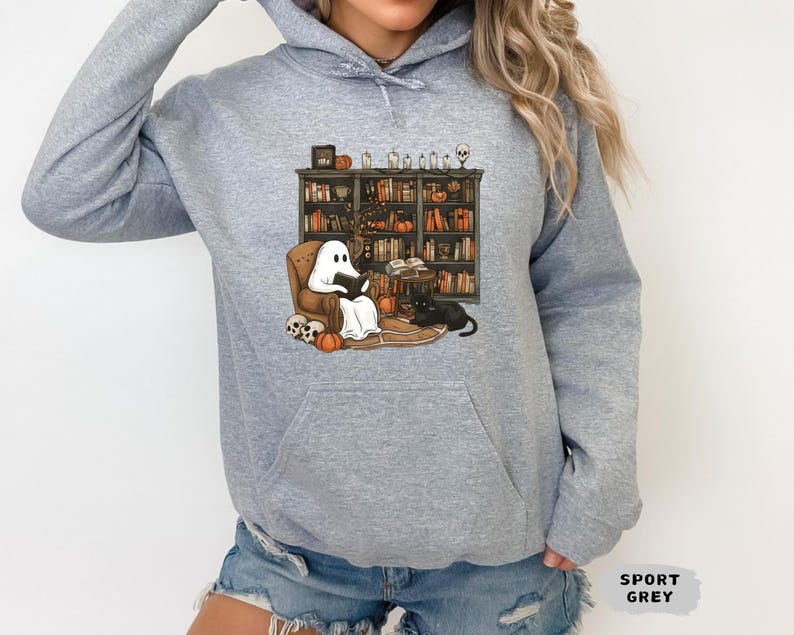 May include: A light gray hoodie featuring a Halloween-themed graphic. The graphic depicts a ghost reading in a chair in front of a bookshelf, with pumpkins, a black cat, and skulls. The text "SPORT GREY" is also visible.