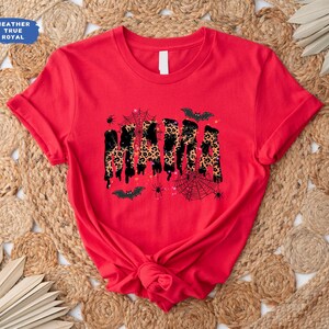 May include: A red t-shirt with the word "MAMA" in a dripping, leopard print design. The shirt features Halloween-themed accents like bats, spiders, and spiderwebs. The shirt is made of a soft material.