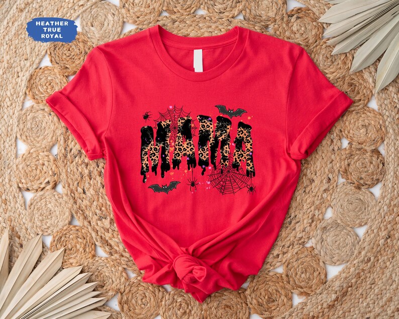 May include: A red t-shirt with the word "MAMA" in a dripping, leopard print design. The shirt features Halloween-themed accents like bats, spiders, and spiderwebs. The shirt is made of a soft material.