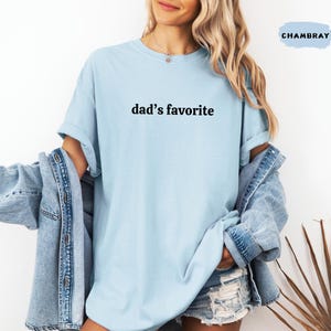 May include: A light blue t-shirt with the text "dad's favorite" in black. The shirt is part of a clothing collection. The model is wearing a denim jacket over the shirt.