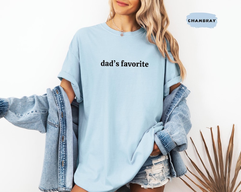 May include: A light blue t-shirt with the text "dad's favorite" in black. The shirt is part of a clothing collection. The model is wearing a denim jacket over the shirt.