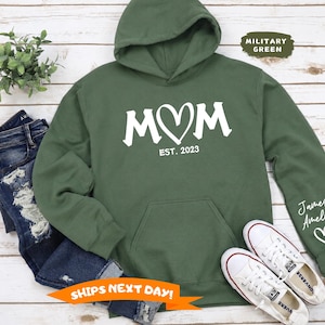 May include: A military green hooded sweatshirt featuring the white text MOM with a heart shape replacing the letter O, and the text EST. 2023 below it. The left sleeve displays the names James and Amelia above a small white heart graphic.