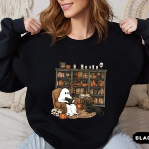 May include: Black sweatshirt featuring a Halloween-themed illustration. The graphic depicts a ghost reading in a cozy armchair in front of a bookshelf, with pumpkins, candles, and a black cat. The word "BLACK" is printed at the bottom.