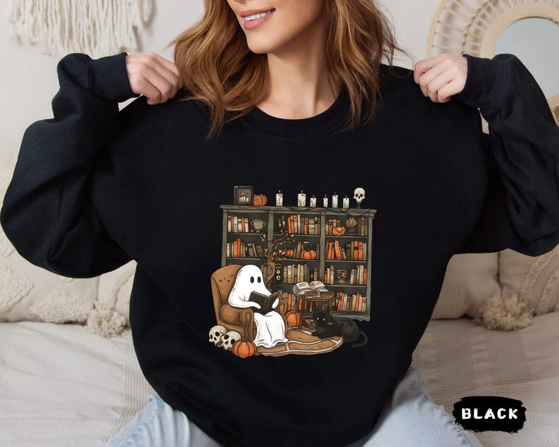 May include: Black sweatshirt featuring a Halloween-themed illustration. The graphic depicts a ghost reading in a cozy armchair in front of a bookshelf, with pumpkins, candles, and a black cat. The word "BLACK" is printed at the bottom.
