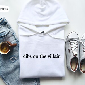 May include: A white hoodie with the text "dibs on the villain" is displayed next to a pair of grey trainers and a pair of blue jeans. A cup of coffee sits on the jeans. The word "WHITE" is in the upper left corner.