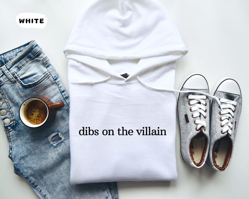 May include: A white hoodie with the text "dibs on the villain" is displayed next to a pair of grey trainers and a pair of blue jeans. A cup of coffee sits on the jeans. The word "WHITE" is in the upper left corner.