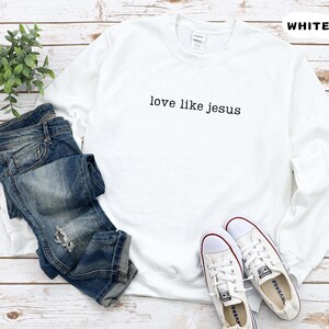 May include: A white sweatshirt with the text "love like jesus" in black, paired with blue jeans and white sneakers. The word "WHITE" is printed in the top right corner. The items are arranged on a light wood surface.