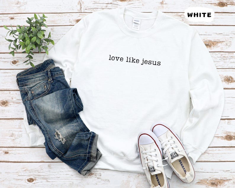 May include: A white sweatshirt with the text "love like jesus" in black, paired with blue jeans and white sneakers. The word "WHITE" is printed in the top right corner. The items are arranged on a light wood surface.
