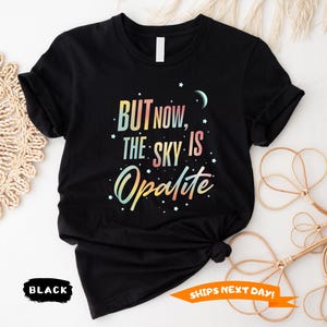 May include: Black t-shirt with the text "BUT NOW, THE SKY IS Opalite" in colorful lettering. The design includes stars and a crescent moon. The shirt is knotted at the bottom.
