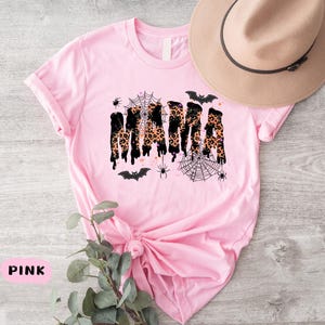 May include: Pink t-shirt with the word "MAMA" in a black and leopard print design, with Halloween-themed spiderwebs, spiders, and bats. A tan hat with a black band rests on the shirt.