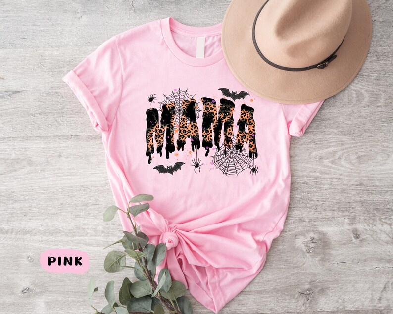 May include: Pink t-shirt with the word "MAMA" in a black and leopard print design, with Halloween-themed spiderwebs, spiders, and bats. A tan hat with a black band rests on the shirt.
