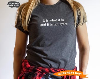 May include: Heather dark grey t-shirt with the text "it is what it is and it is not great" in white. The shirt is a crew neck style. A red and black plaid shirt is tied around the waist.