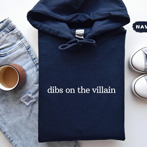 May include: A navy blue hoodie with the text "dibs on the villain" in white. The image also shows light blue jeans, a brown mug, and grey trainers.